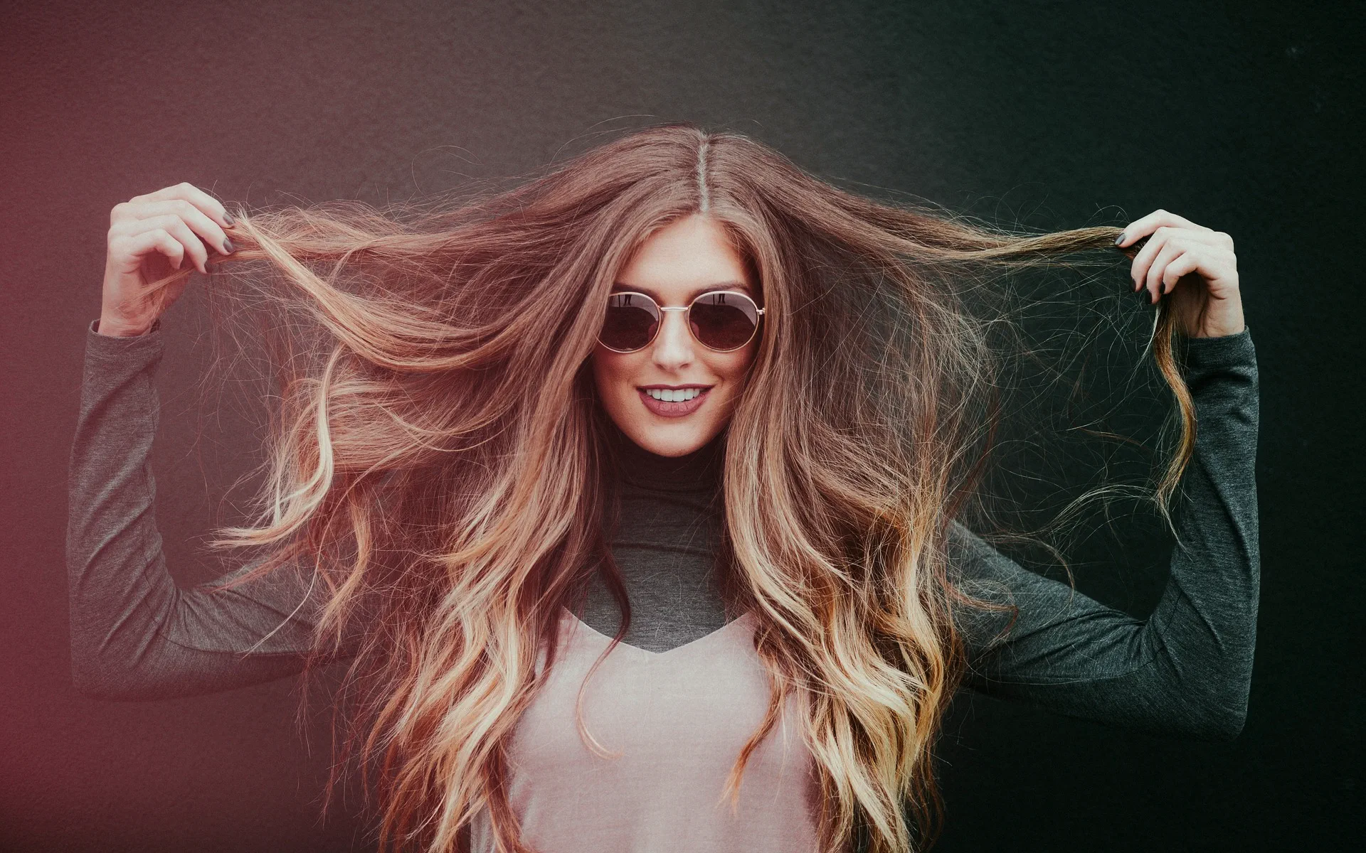 A smiling woman wearing sunglasses, playfully pulling on her hair strands—showcasing confidence and breaking free from people-pleasing.